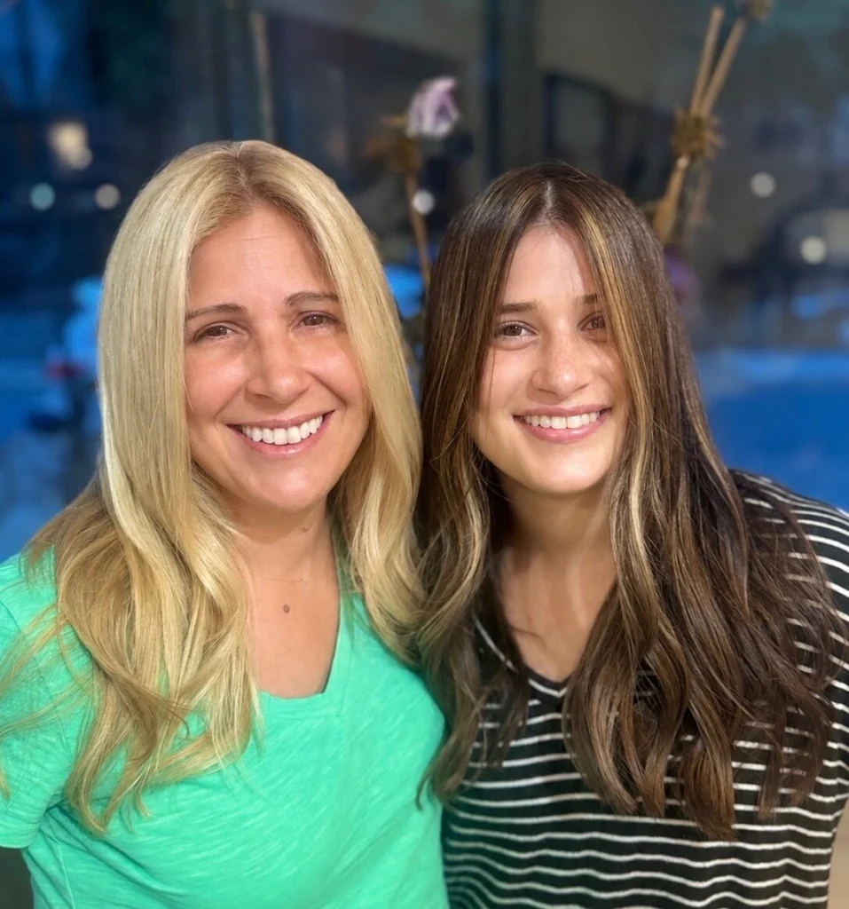 Two smiling clients: one with long golden blonde hair, one with long brunette hair and face-framing highlights